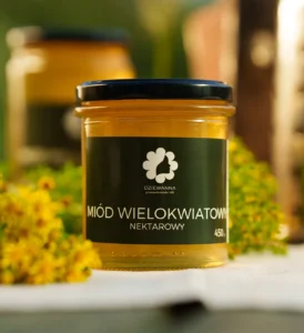 słoik miodu wielokwiatowego Dziewanna na jasnej powierzchni, obok żółte kwiaty i drugi słoik w tle
